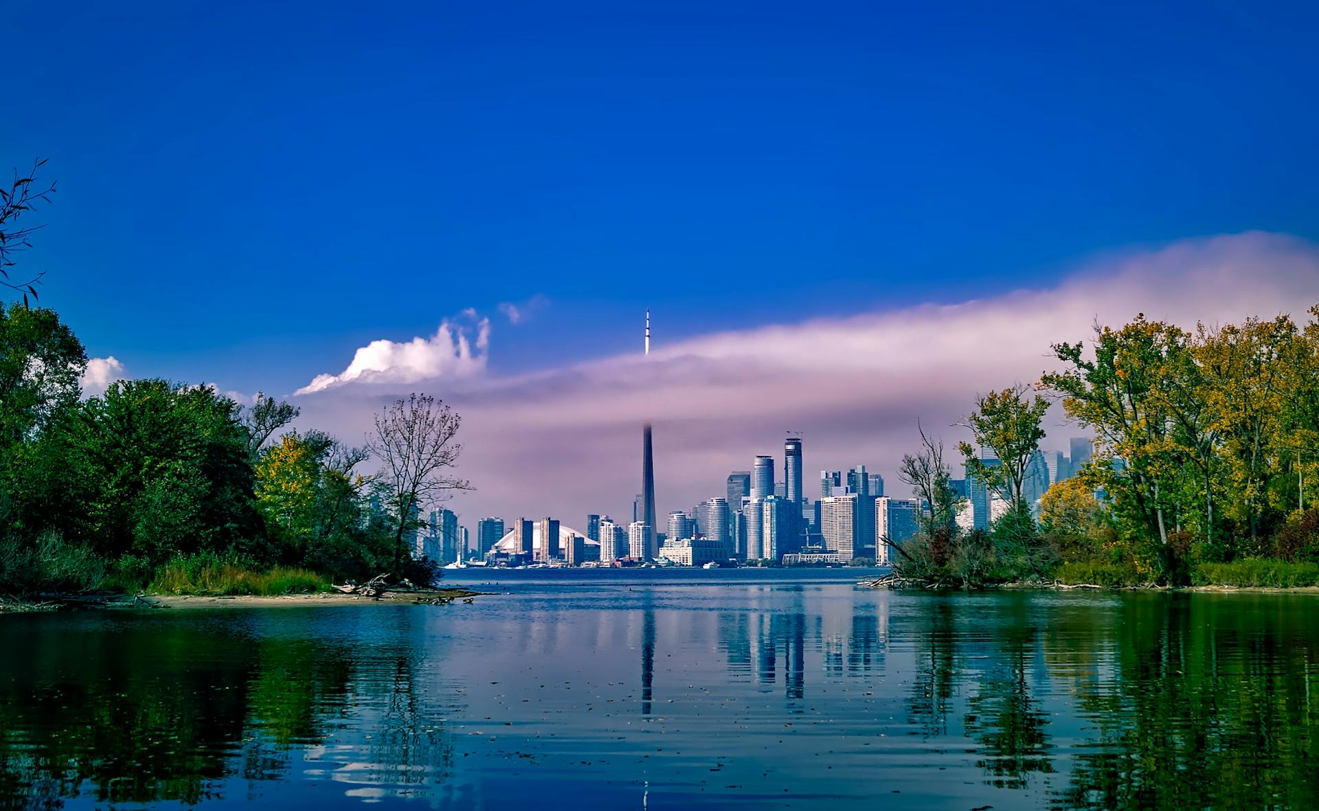 Scenic view of Toronto skyline across a reflective lake with lush greenery and clear blue sky.