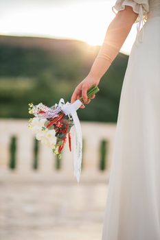 A bride in a white dress holds a beautiful bouquet with a scenic outdoor backdrop during sunset.