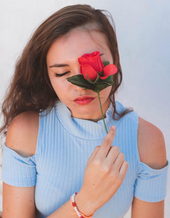Free High angle of tranquil young female in casual clothes covering closed eye with fresh red rose against white background Stock Photo