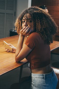 A young woman with curly hair casually sitting indoors holding a smartphone and looking back.