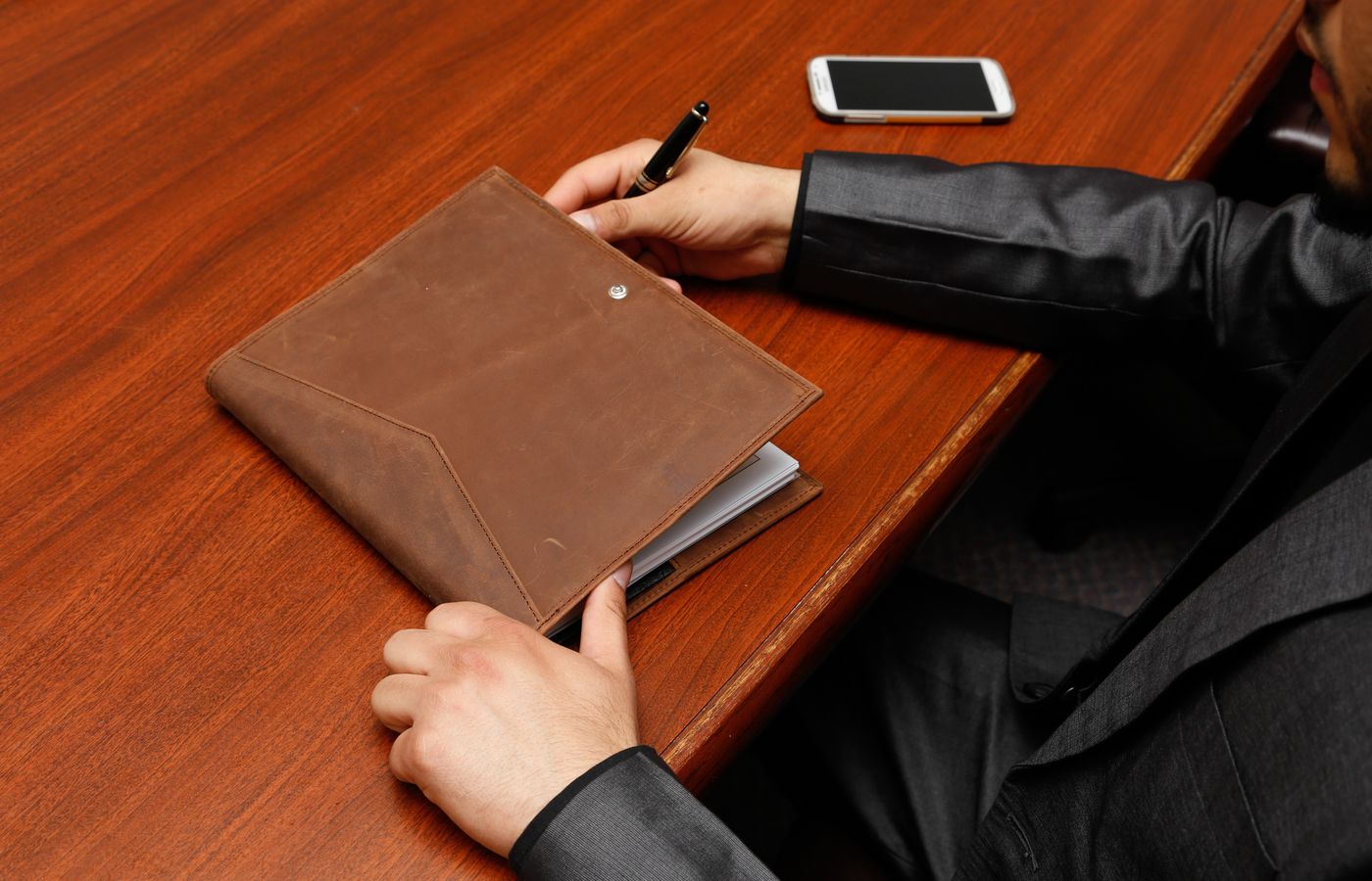 Male executive writing notes during a business meeting using a leather notebook.