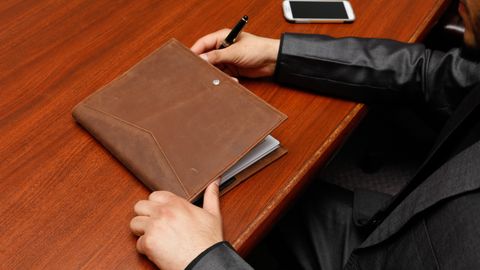 Male executive writing notes during a business meeting using a leather notebook.