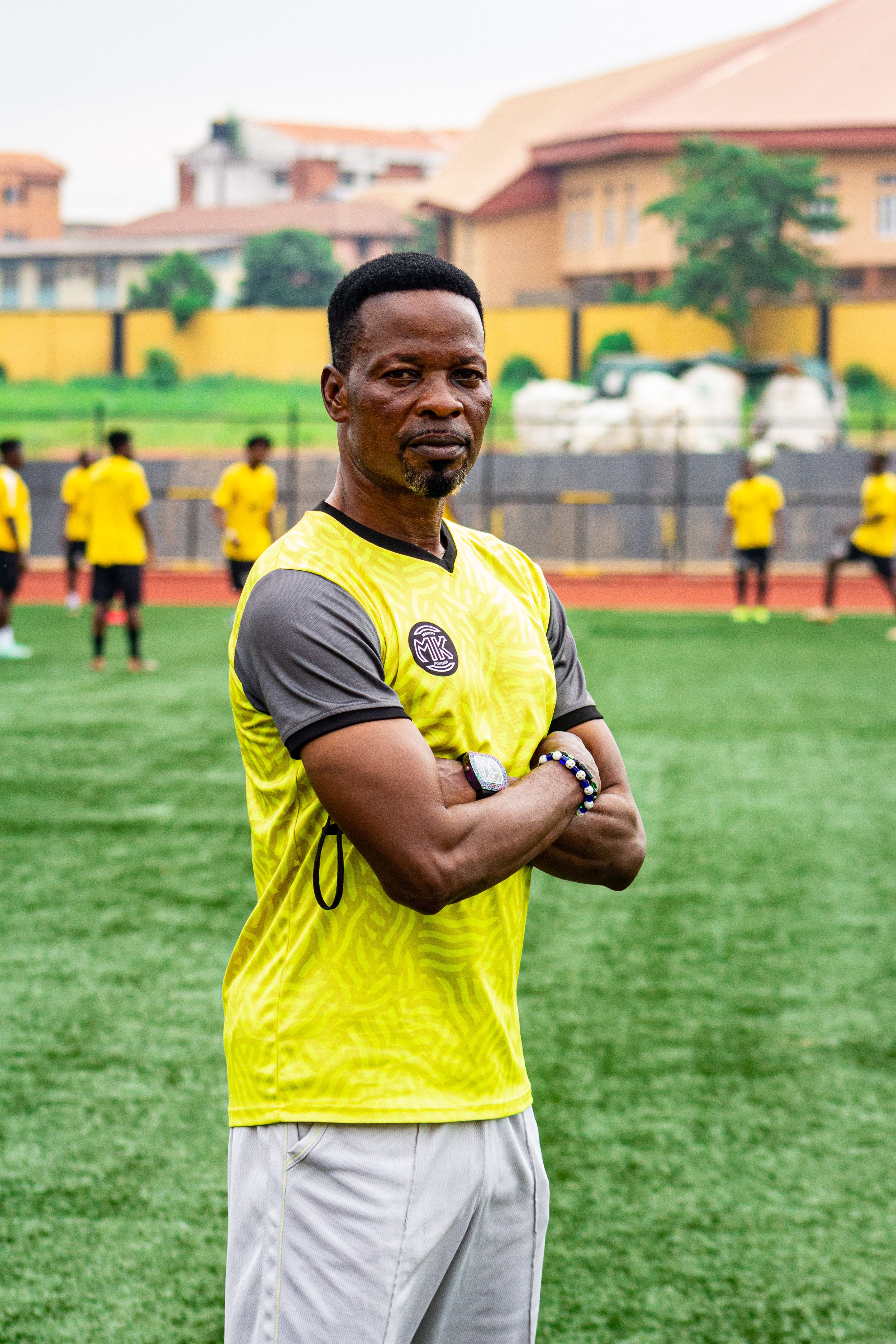 A confident soccer coach with arms crossed on a football field, surrounded by players training.