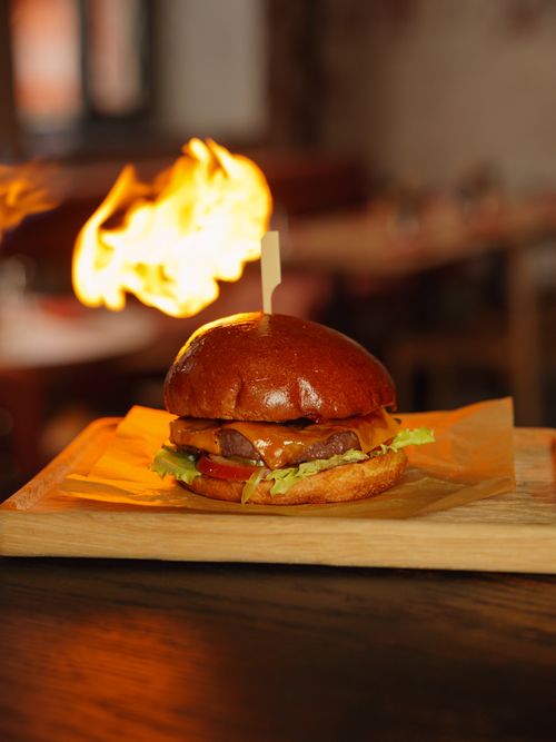 Free Tasty gourmet burger served on a wooden tray with a dramatic flame in a restaurant setting. Stock Photo