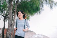 一位穿着休闲服的亚洲女人在海边的树旁微笑摆姿，洋溢着幸福