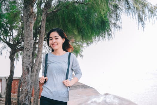 一位穿着休闲服的亚洲女人在海边的树旁微笑摆姿，洋溢着幸福