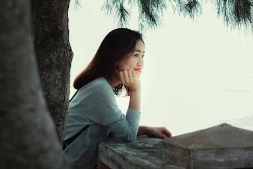 Free Photo of Smiling Woman Standing Near Tree Leaning on Stone Railing Looking Out into the Distance Stock Photo