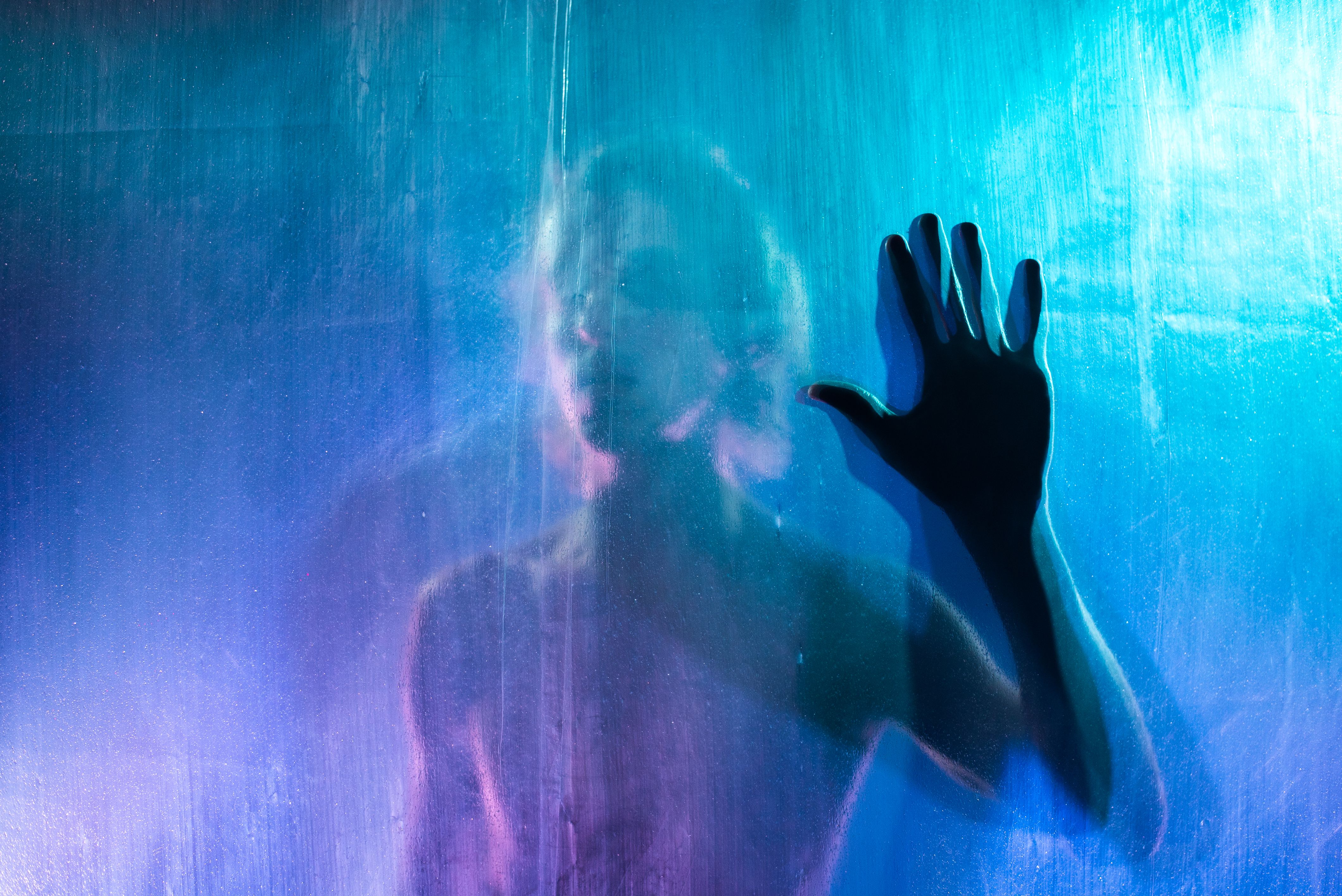 Mysterious silhouette of a person behind a wet transparent surface with a blue light in background.