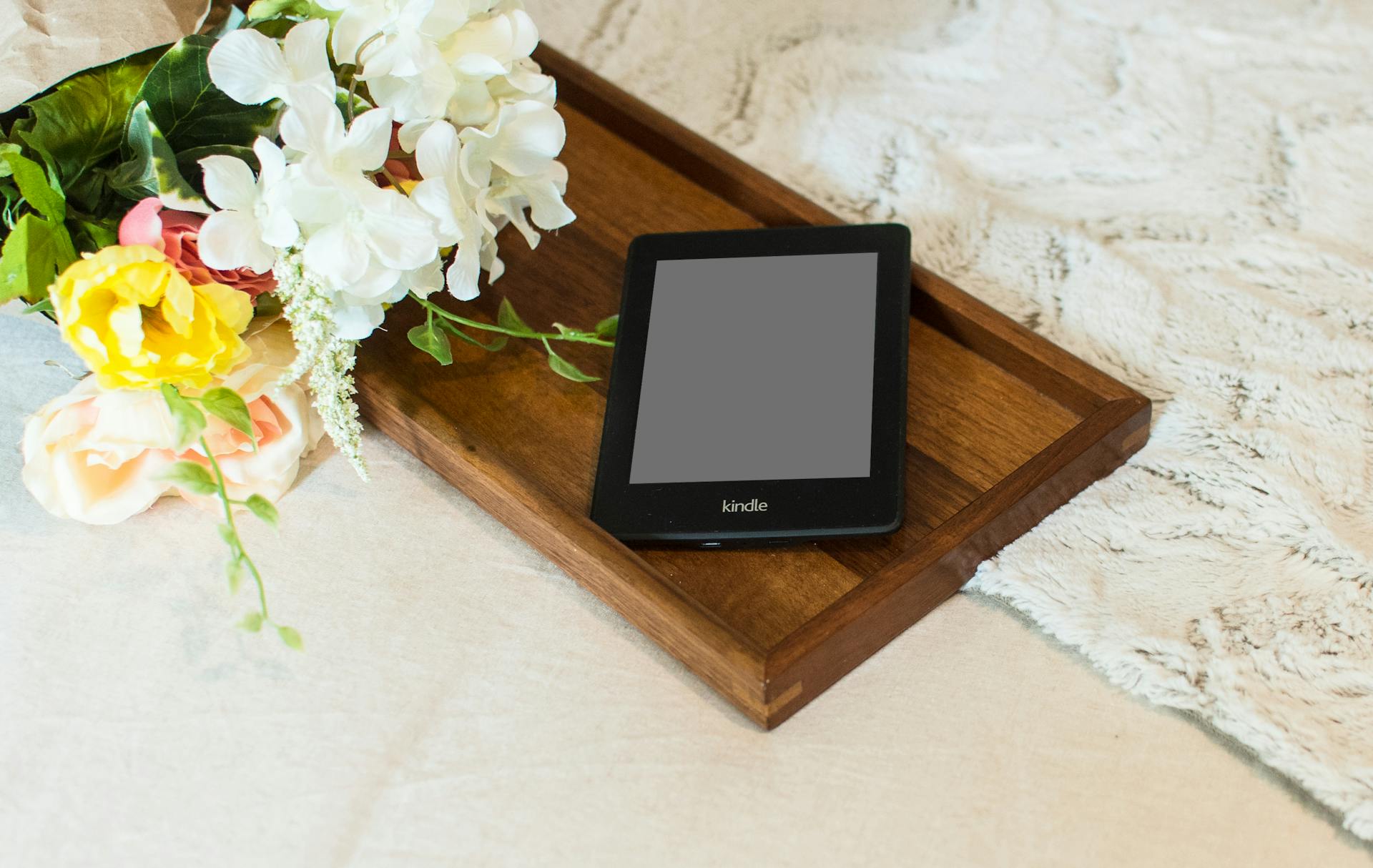 A Kindle e-reader resting on a wooden tray beside vibrant artificial flowers, creating a cozy and inviting scene.