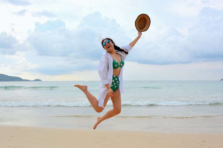 Woman Jumping On Seashore And Holding Hat