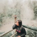 一名黑衣女子倚着竹栏杆，俯瞰雾气缭绕的温泉，放松身心