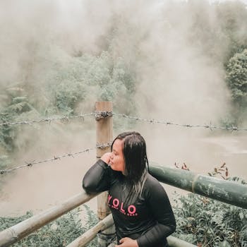 一名黑衣女子倚着竹栏杆，俯瞰雾气缭绕的温泉，放松身心