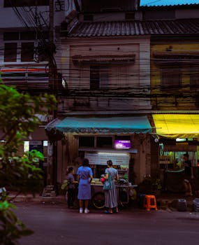 People gather at a food stall on a dimly lit urban street during twilight.