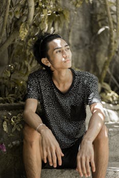 年轻人穿着休闲服，坐在户外的台阶上，在斑驳的阳光下显得若有所思