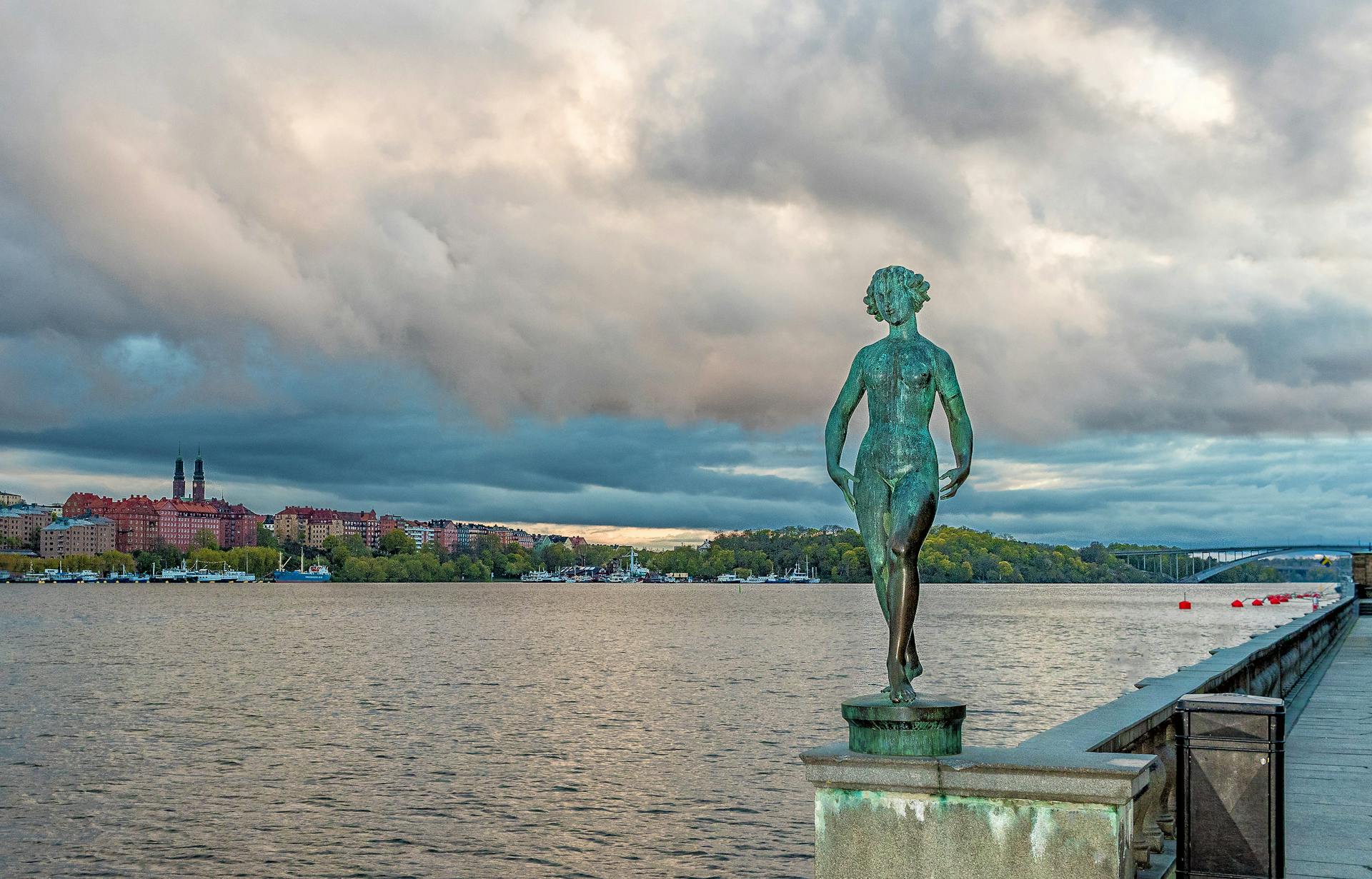 Scenic view of a statue by the Stockholm waterfront with cityscape and dramatic clouds.