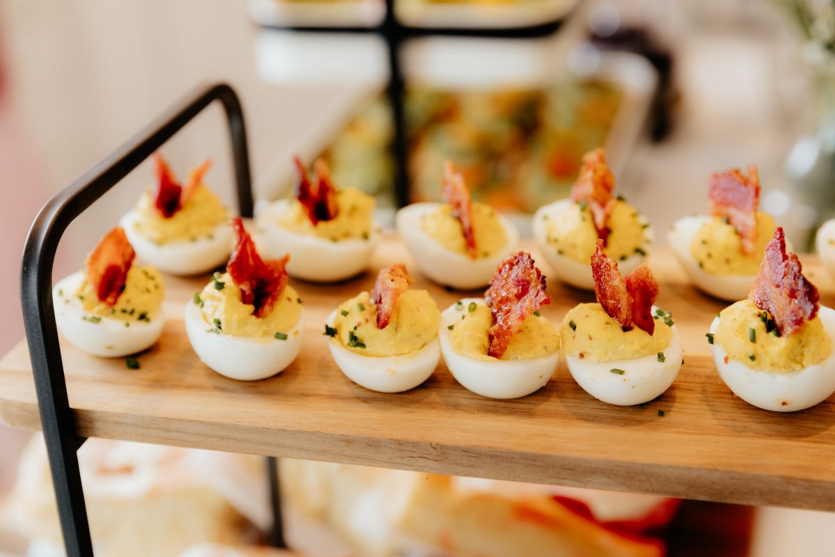 Deviled eggs topped with crisp bacon pieces and chives lined up on a wooden serving tier