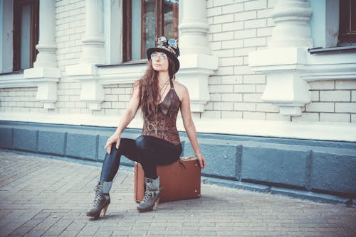 女人坐在大樓前的棕色手提箱上的照片