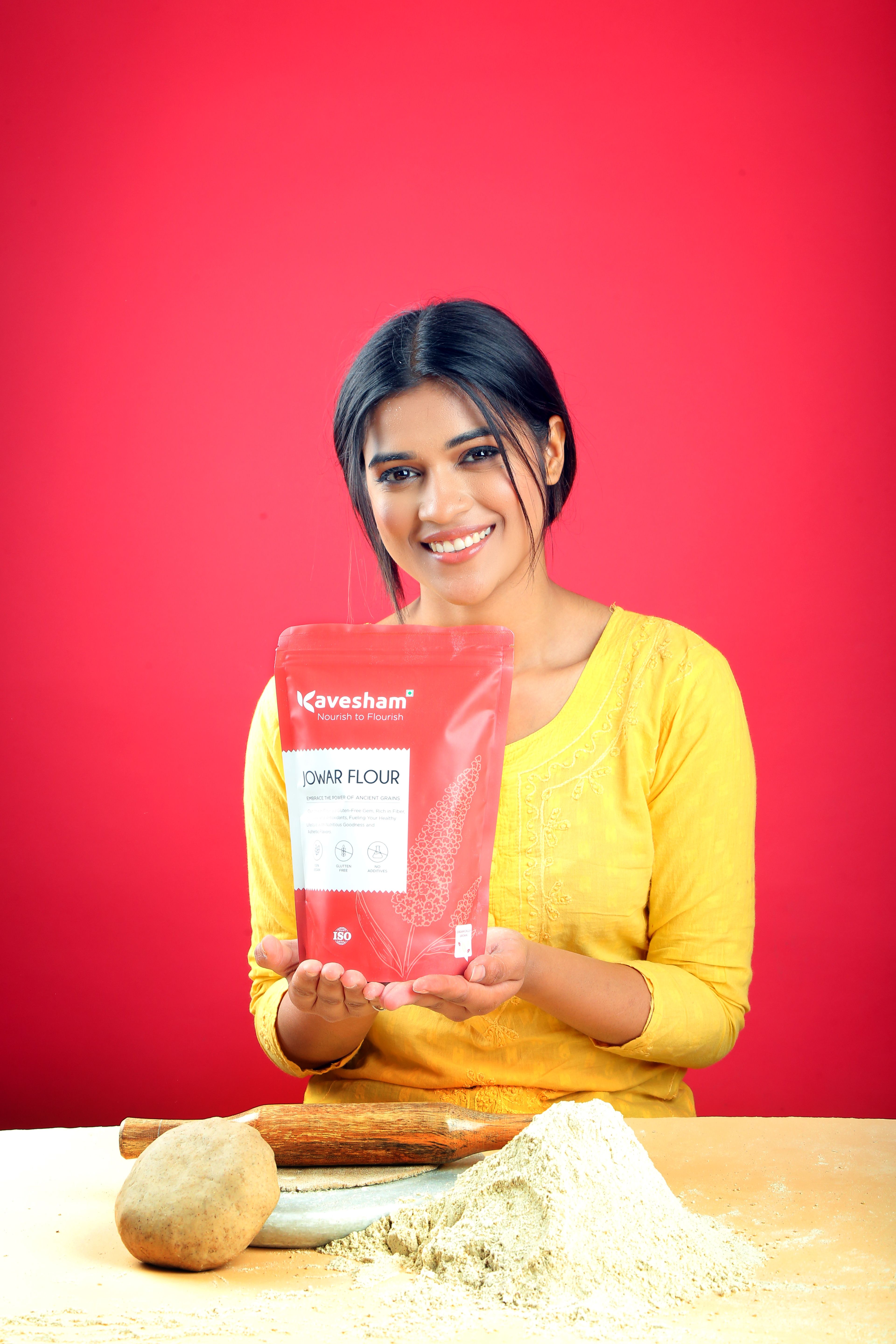 An Indian woman in a bright kitchen presenting a bag of flour with baking ingredients, representing egg-free baking preparation.