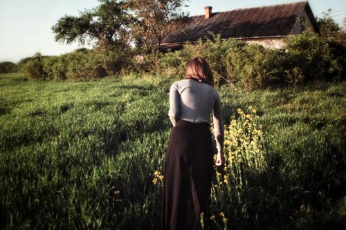 Free A woman walks through a lush green field towards an old house under a clear sky. Stock Photo