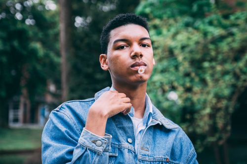 Free Close-up portrait of a young man outdoors with daisy in mouth, wearing denim. Stock Photo