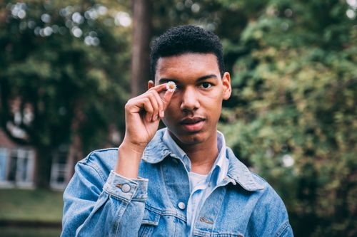 Free Portrait of a young man outdoors holding a daisy flower up to his eye, wearing a denim jacket. Stock Photo