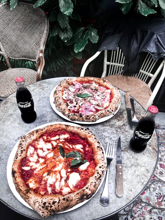 Two Pizzas on Plates, Forks and Knives and Coca Cola Sodas on top of a Table