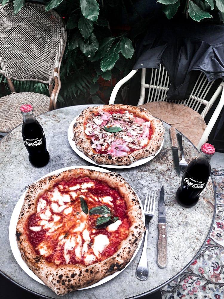 Two Pizzas On Plates, Forks And Knives And Coca Cola Sodas On Top Of A Table