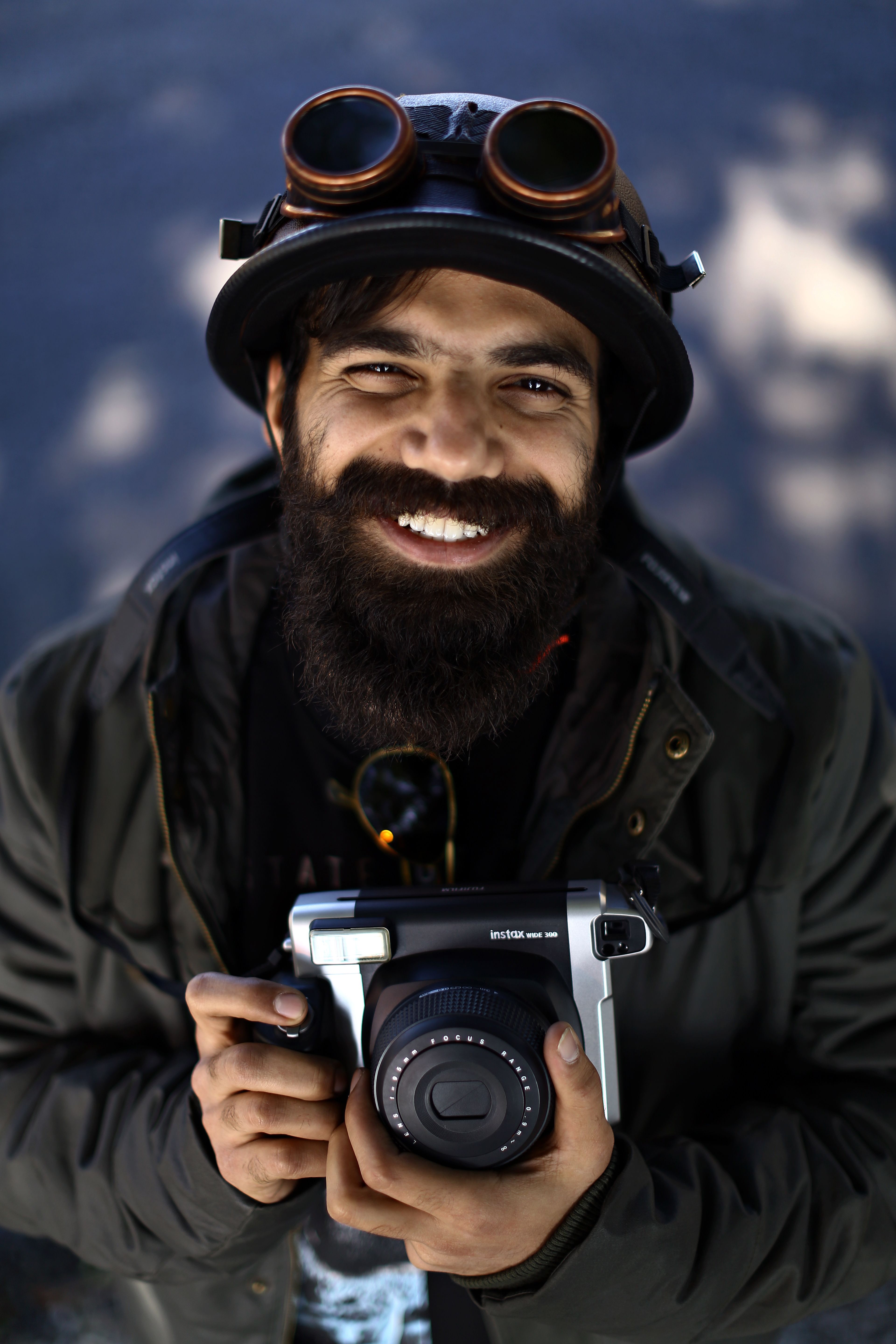 A smiling man wearing goggles holds a vintage camera with a joyful expression.