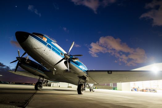 A classic propeller airplane on a runway at twilight with colorful clouds and hangar in the background.