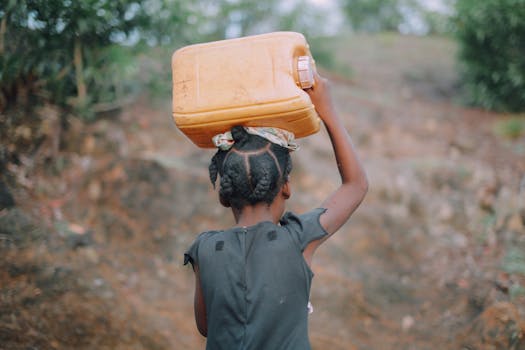 A young girl with braided hair carries a jug of water on her head in a rural area of Haiti.