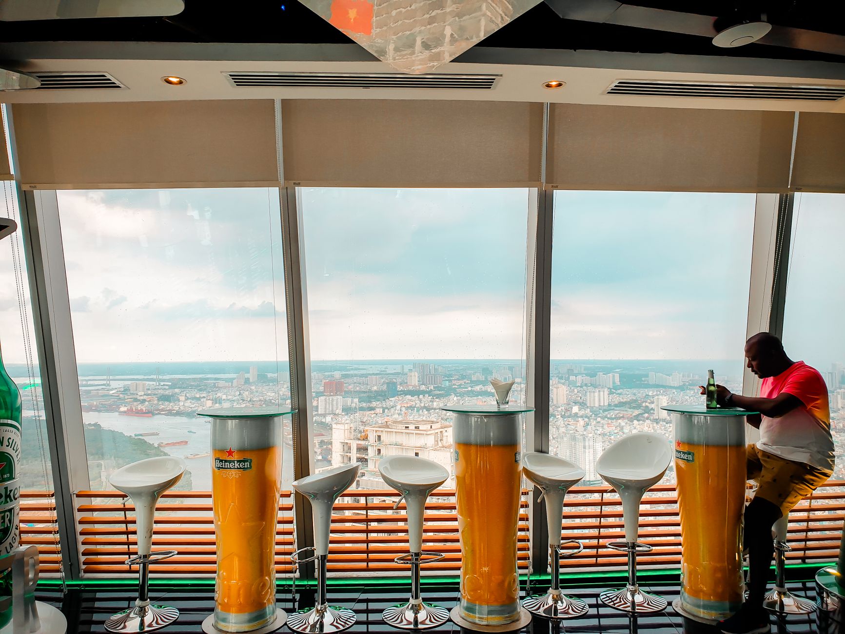 Man sitting at a rooftop bar, enjoying a drink with a cityscape view through large windows.