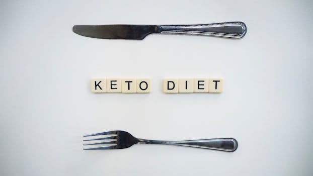 Top view of a knife, fork, and scrabble tiles spelling 'Keto Diet' on a white surface.