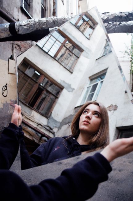 A woman holds a mirror reflecting a building facade, creating a unique urban portrait.