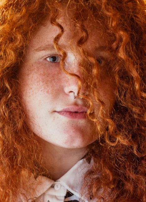Close-up portrait of a woman with vibrant red curly hair and freckles, wearing a septum ring.