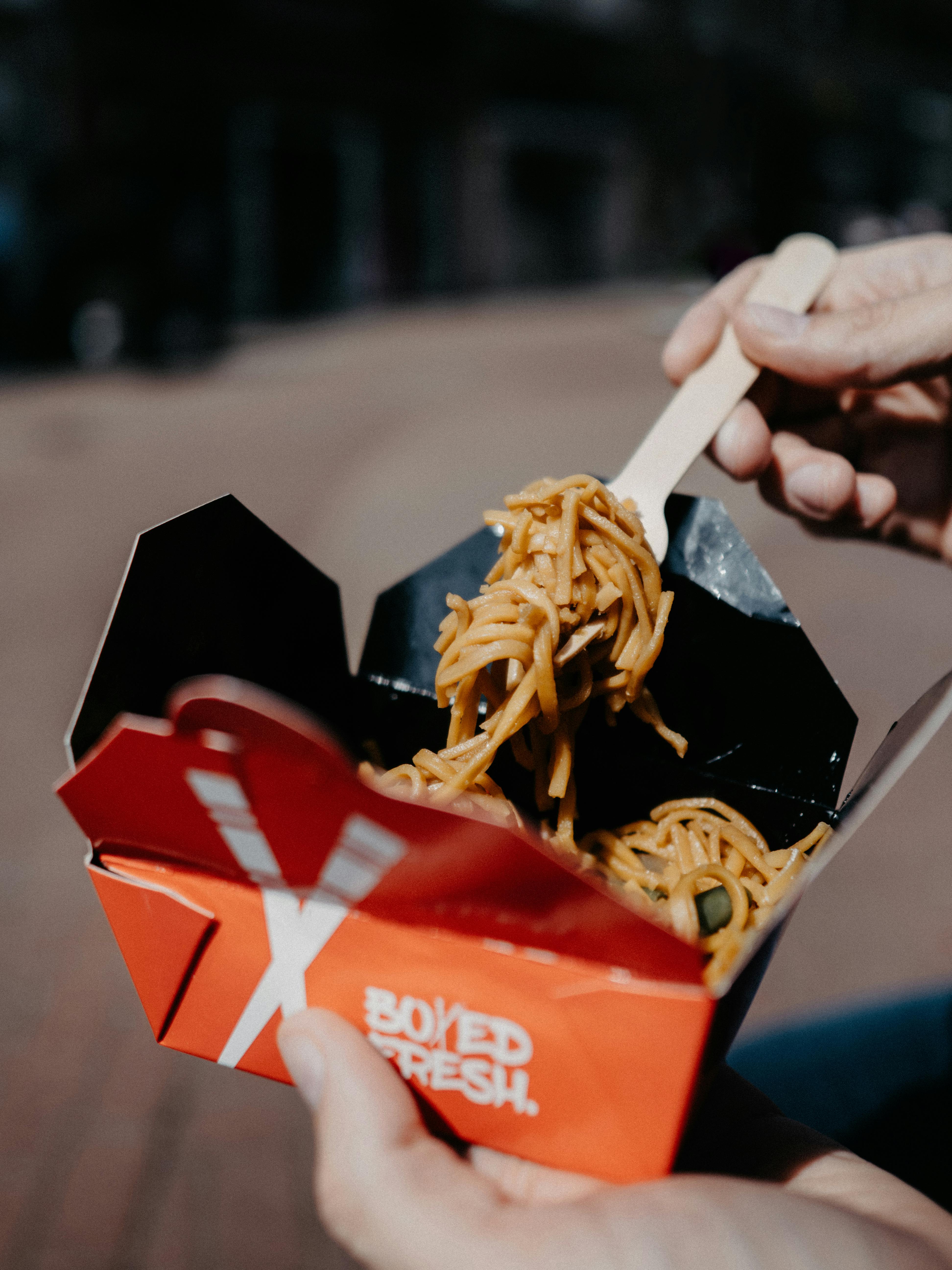 Close-up of delicious street food noodles in a red box outdoors, hand holding a wooden fork.