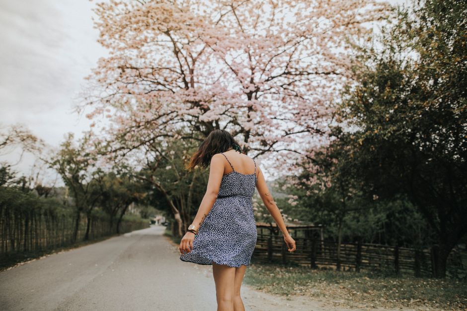A young woman in a dress walks on a tree-lined road during spring, embracing nature and tranquility.