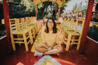 一个开朗的女人穿着黄色连衣裙，坐在色彩缤纷的木船上，周围是充满活力的椅子，沐浴在日光下