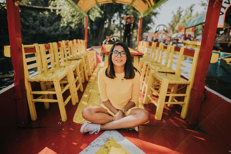 A cheerful woman in a yellow dress sits on a colorful wooden boat surrounded by vibrant chairs in daylight.