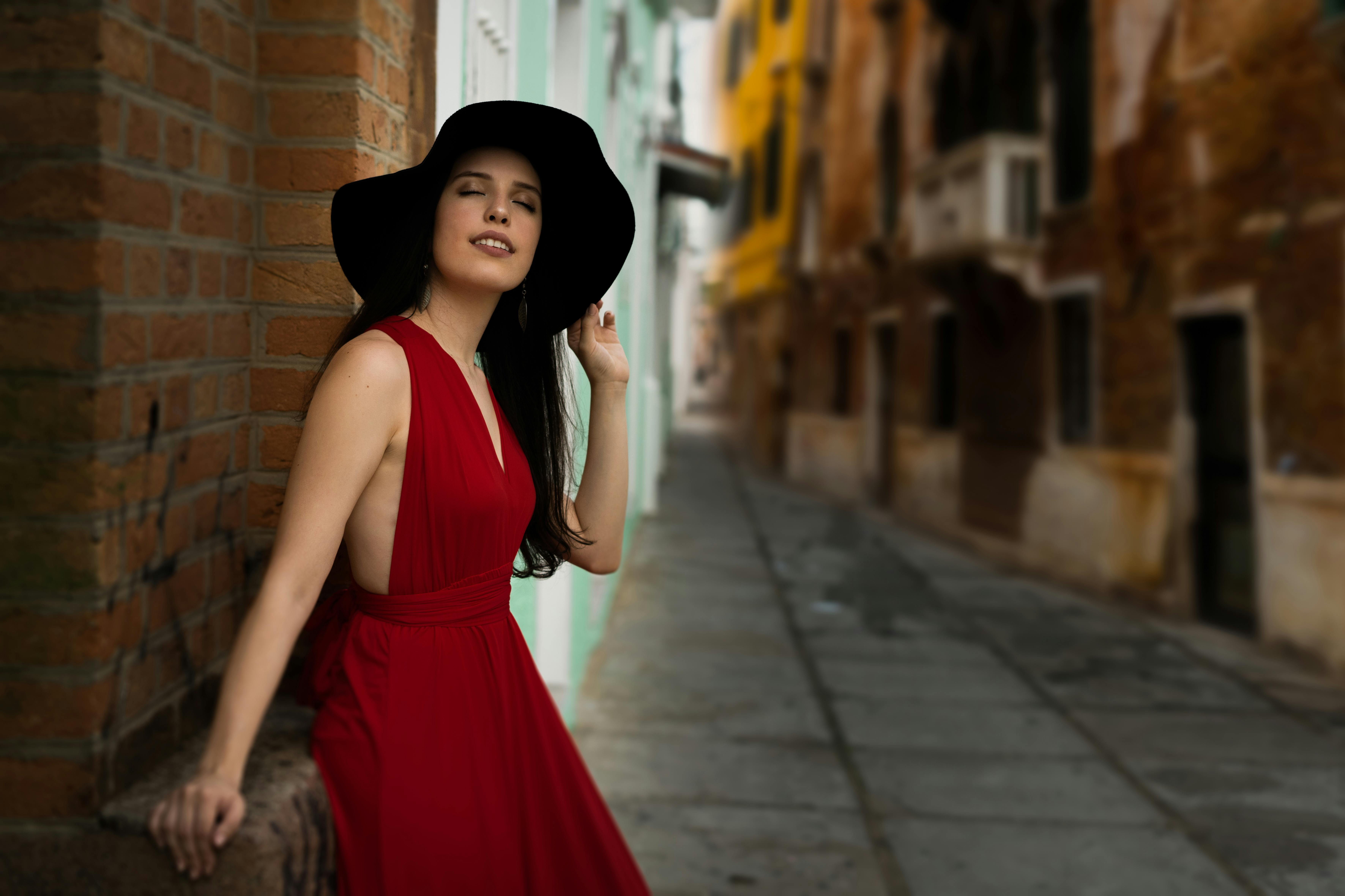 Photo of Woman With Her Eyes Closed Wearing a Red Dress and Black Hat Leaning on Red Brick Wall in Alley