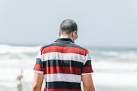 一个穿着条纹衬衫的男人，站在晴朗天空下的沙滩上，面朝大海