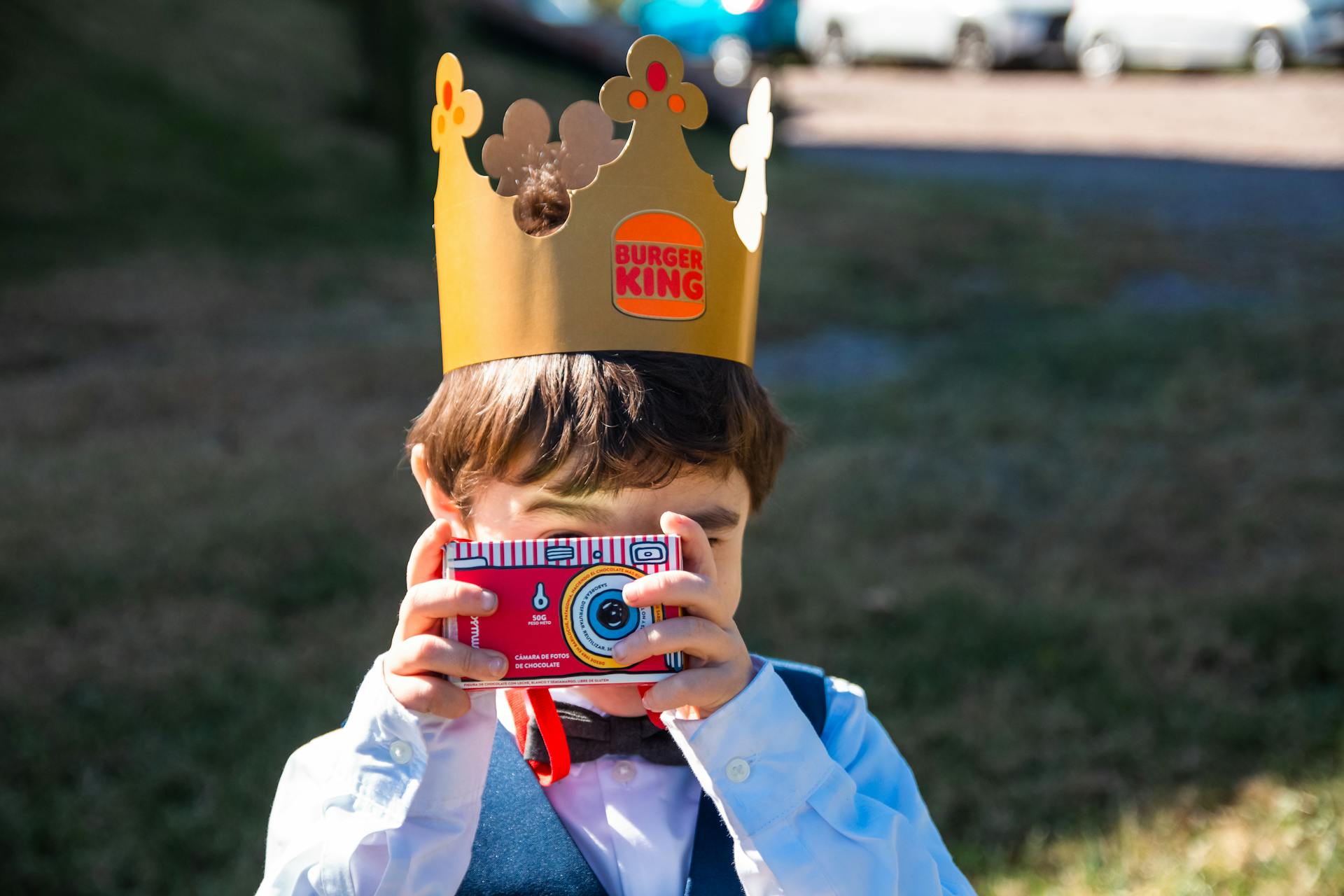 Young boy with Burger King crown using paper camera outside on a sunny day.