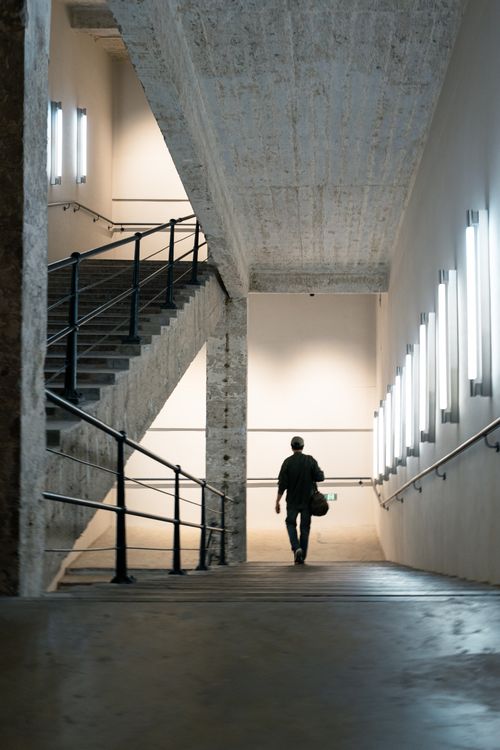 Free A lone man walking down a dimly lit concrete staircase in an industrial building interior. Stock Photo