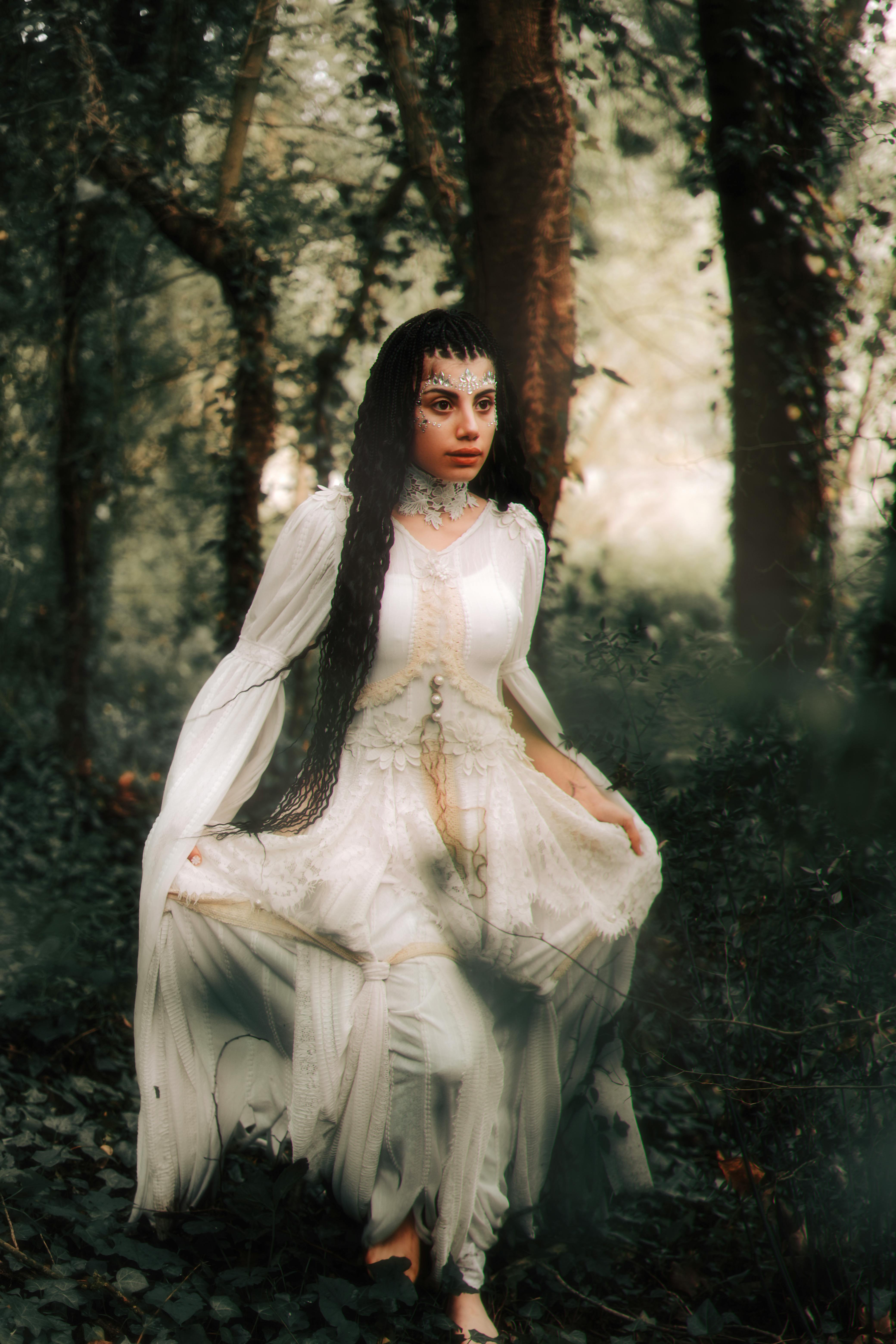 Woman with long hair and creative makeup in a white dress posing in a mystical forest.