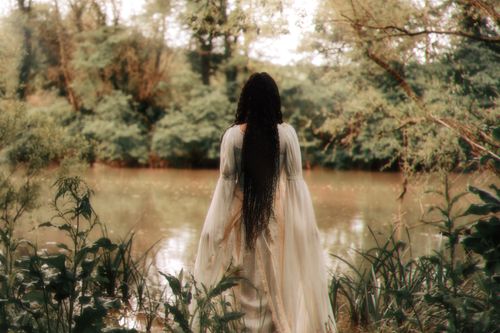 Free Brunette woman with long braids standing by a peaceful river in nature. Stock Photo