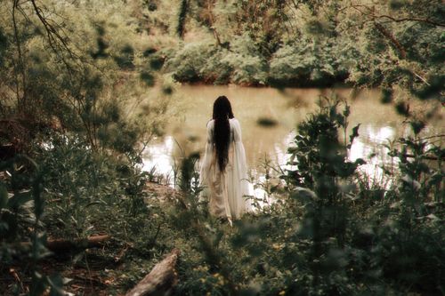 Free A serene scene of a woman in a white dress standing by a riverbank surrounded by lush greenery. Stock Photo