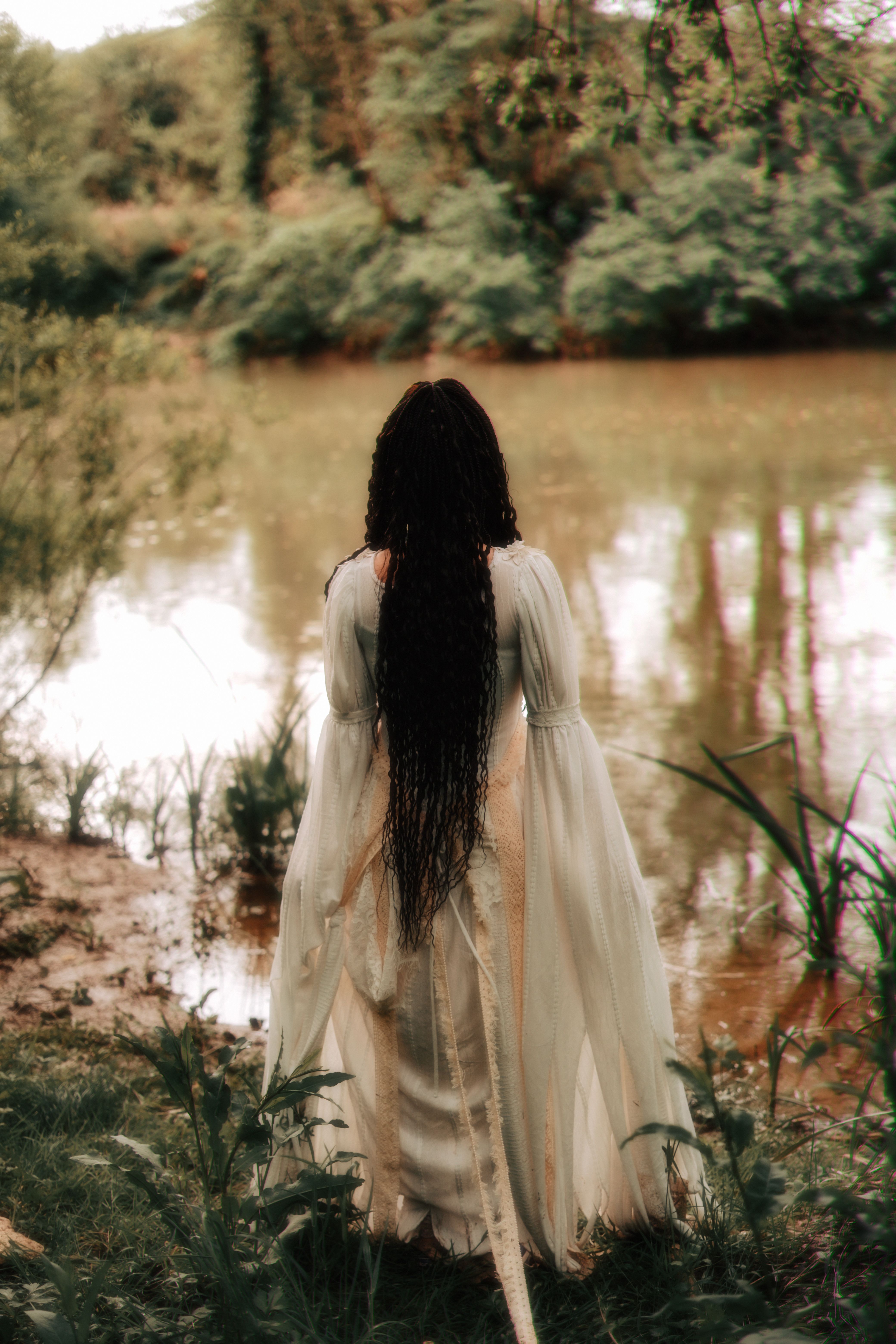 A woman in a white dress with long hair stands by a tranquil riverbank, embracing nature.