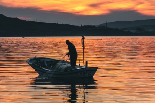 Free A lone fisherman on a small boat silhouetted by a stunning sunset over calm waters. Stock Photo