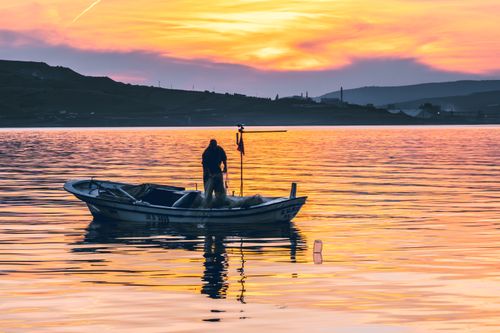 Free A lone fisherman stands on a boat during a vibrant sunset over a serene lake. Stock Photo