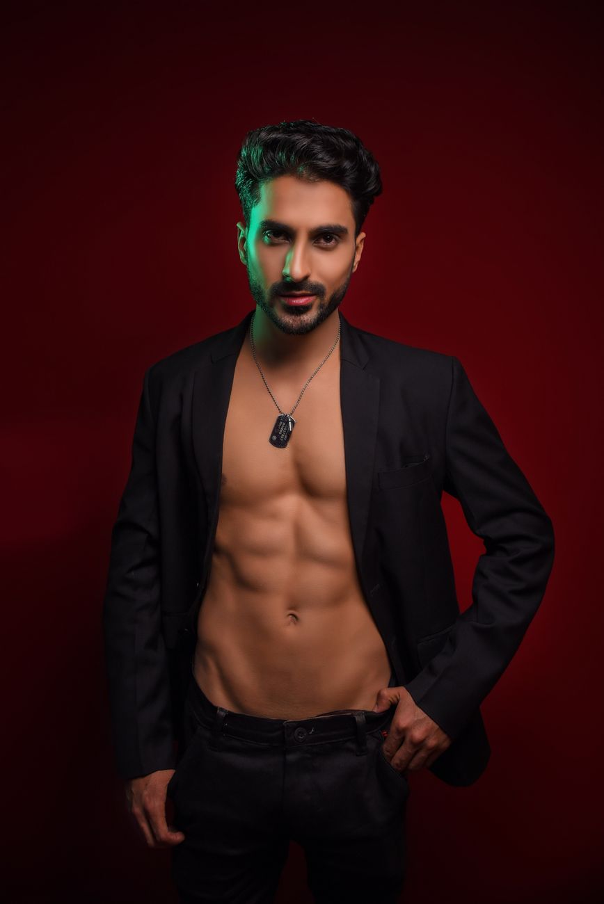 A shirtless man in a black blazer poses confidently against a red background.