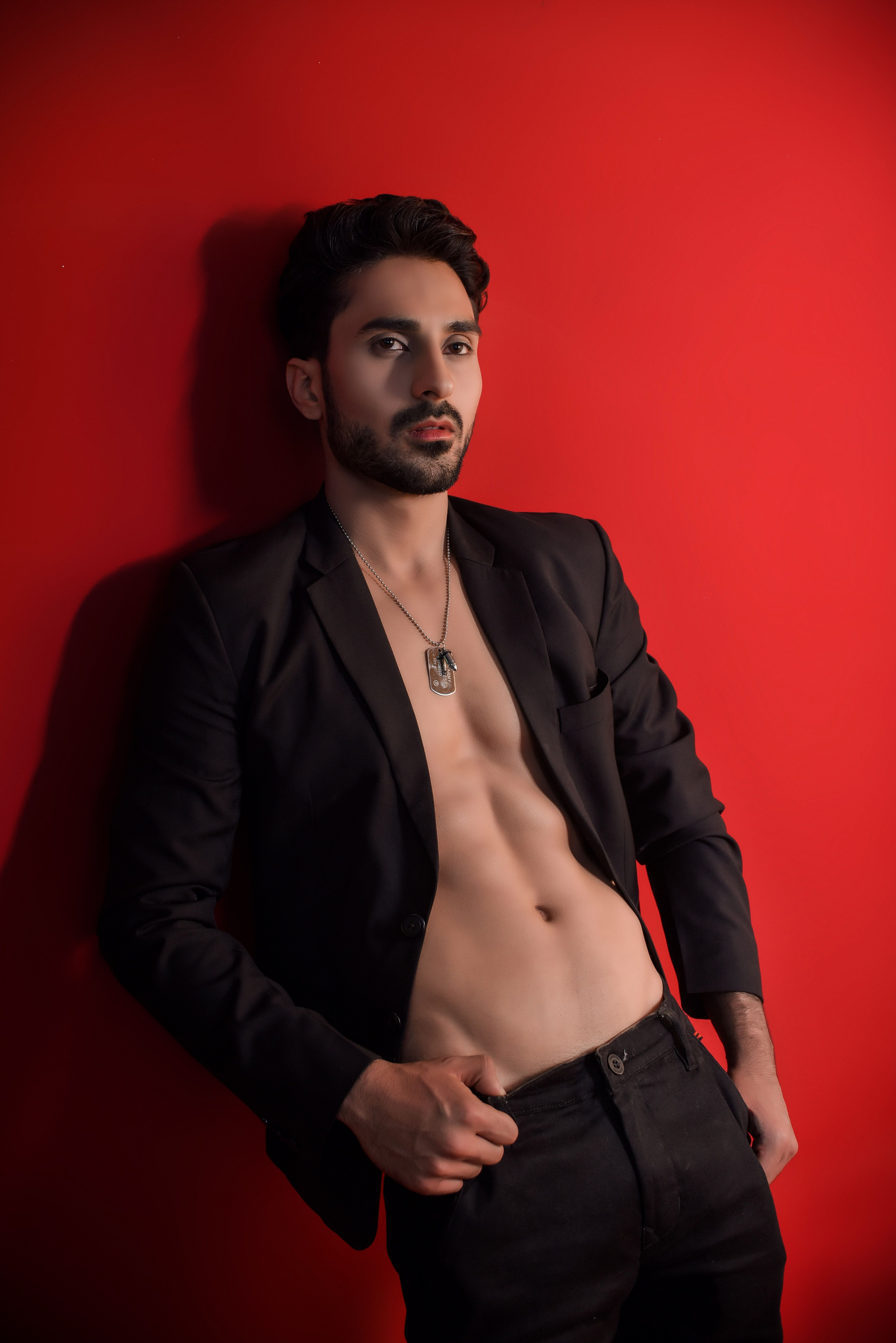 Confident man in a black blazer poses against a striking red wall, showcasing modern style.